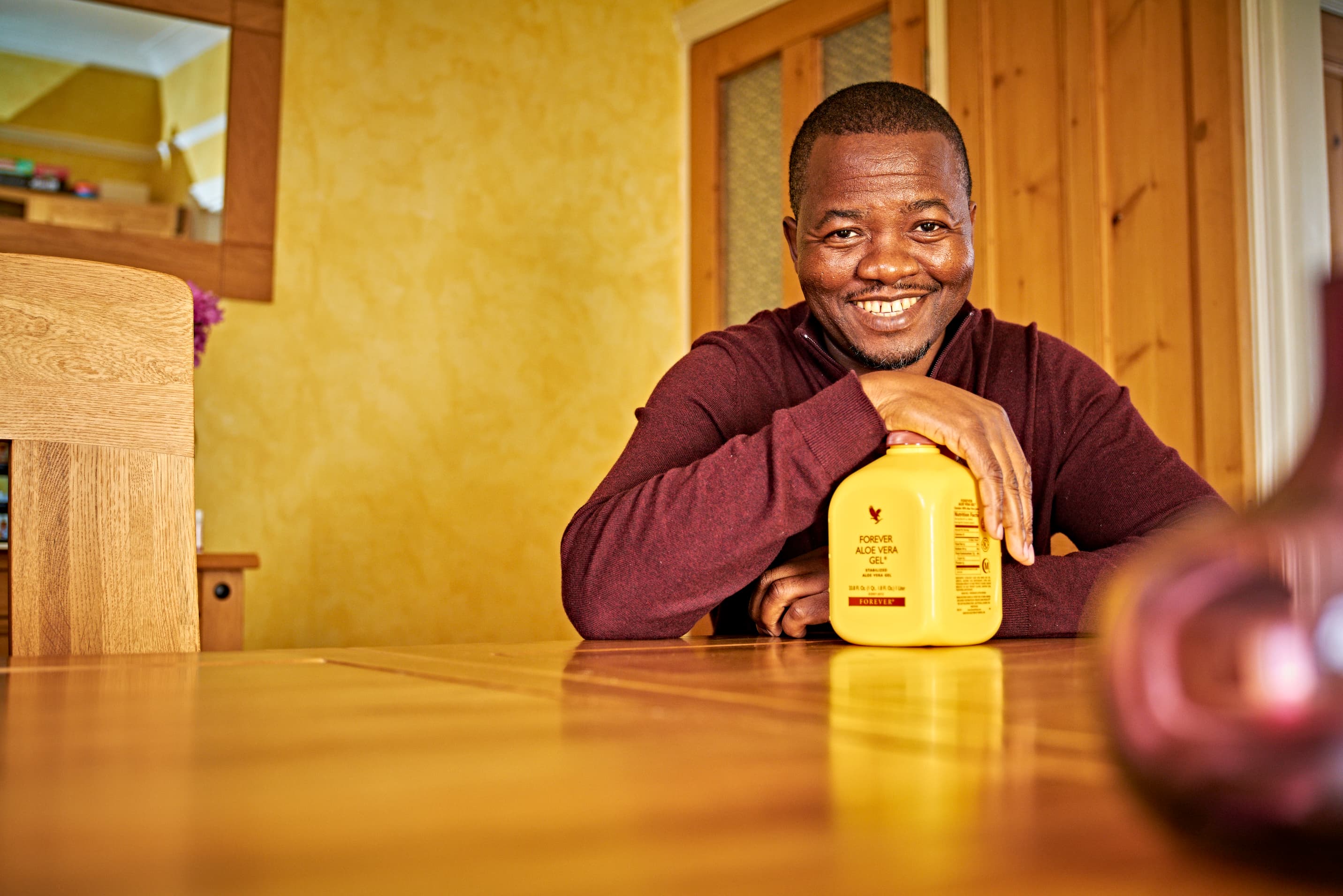 Long-term daily aloe drinker smiling comfortably at home dining table with the iconic yellow Forever Aloe Vera Gel jug